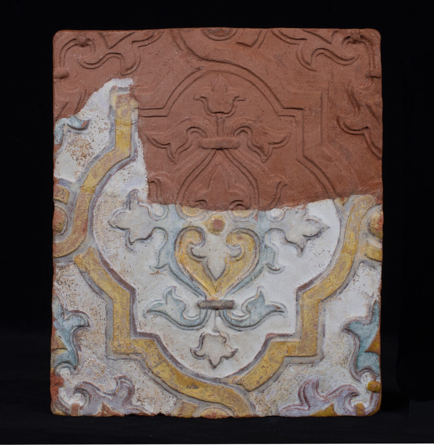 Fragment kafla ceramicznego z częściowo odsłoniętą, barwną dekoracją roślinną w stylu historycznym. W górnej części widoczna surowa czerwona glina z wytłoczonym ornamentem.
