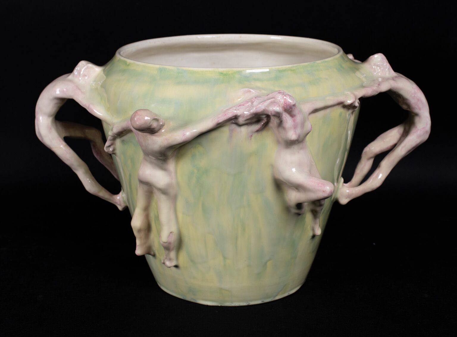 Ceramika w formie dużego naczynia o jasnozielonej powierzchni, ozdobiona figurkami nagich ludzkich postaci, które trzymają się za ręce i tworzą uchwyty oraz zdobią ścianki.