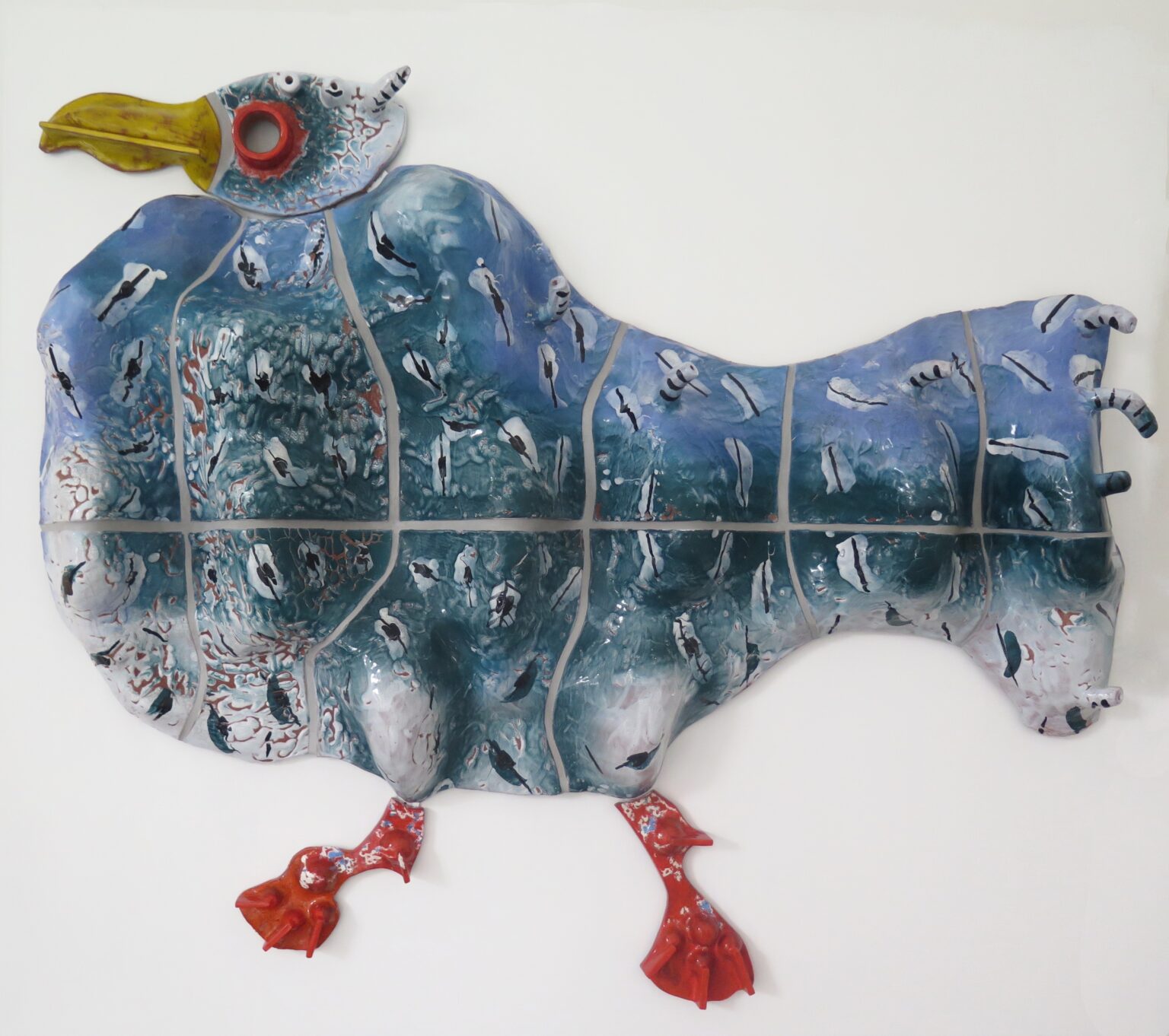 Artystyczna ceramiczna płaskorzeźba w kształcie stylizowanego ptaka, z korpusem pokrytym niebiesko-zielonym szkliwem i malowanymi sylwetkami ptaków. Głowa jest żółta z czerwonym okiem, a nogi intensywnie czerwone.