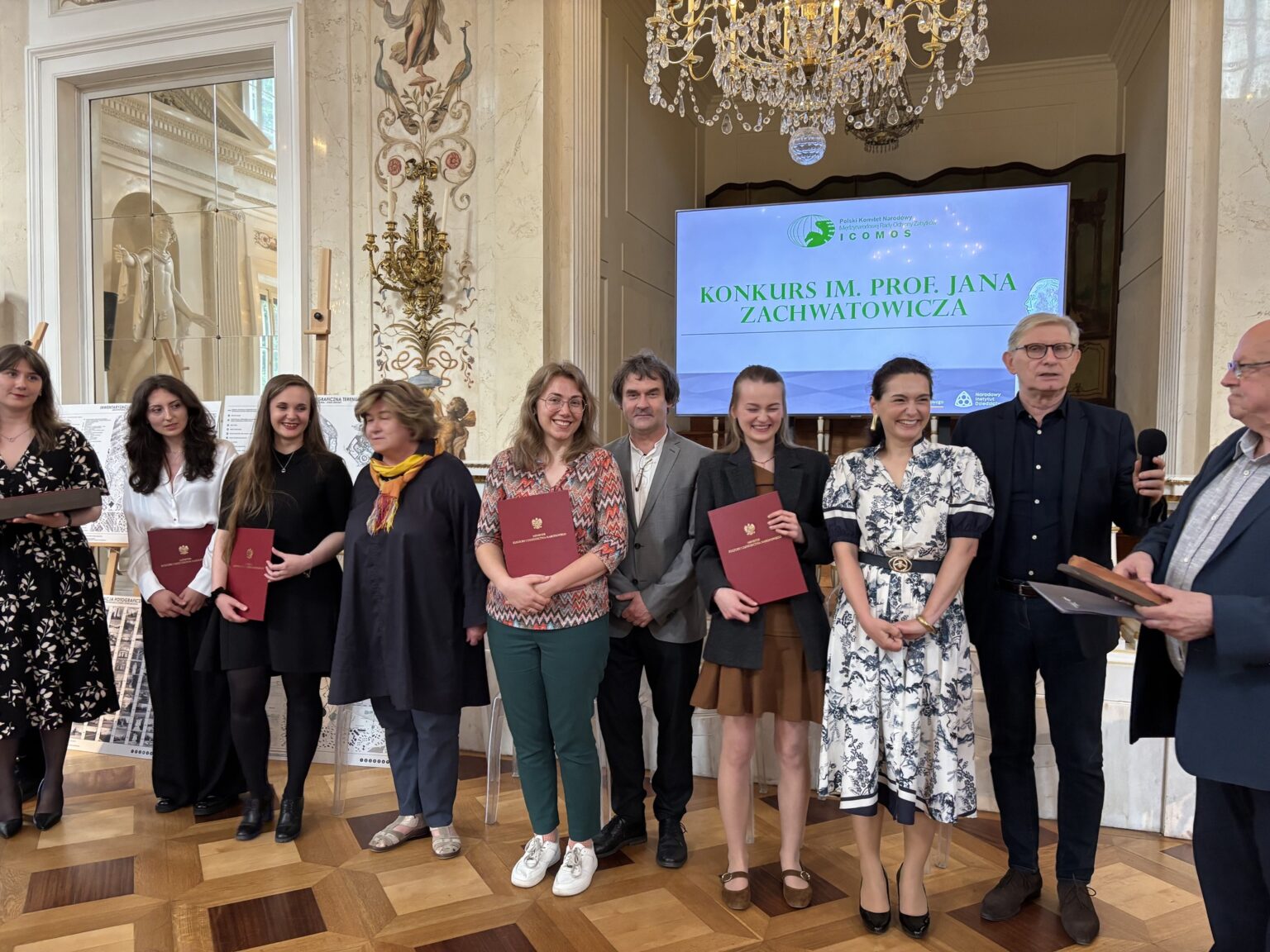 Grupowe zdjęcie laureatów i organizatorów Konkursu im. prof. Jana Zachwatowicza w eleganckim wnętrzu z żyrandolem i ekranem w tle.