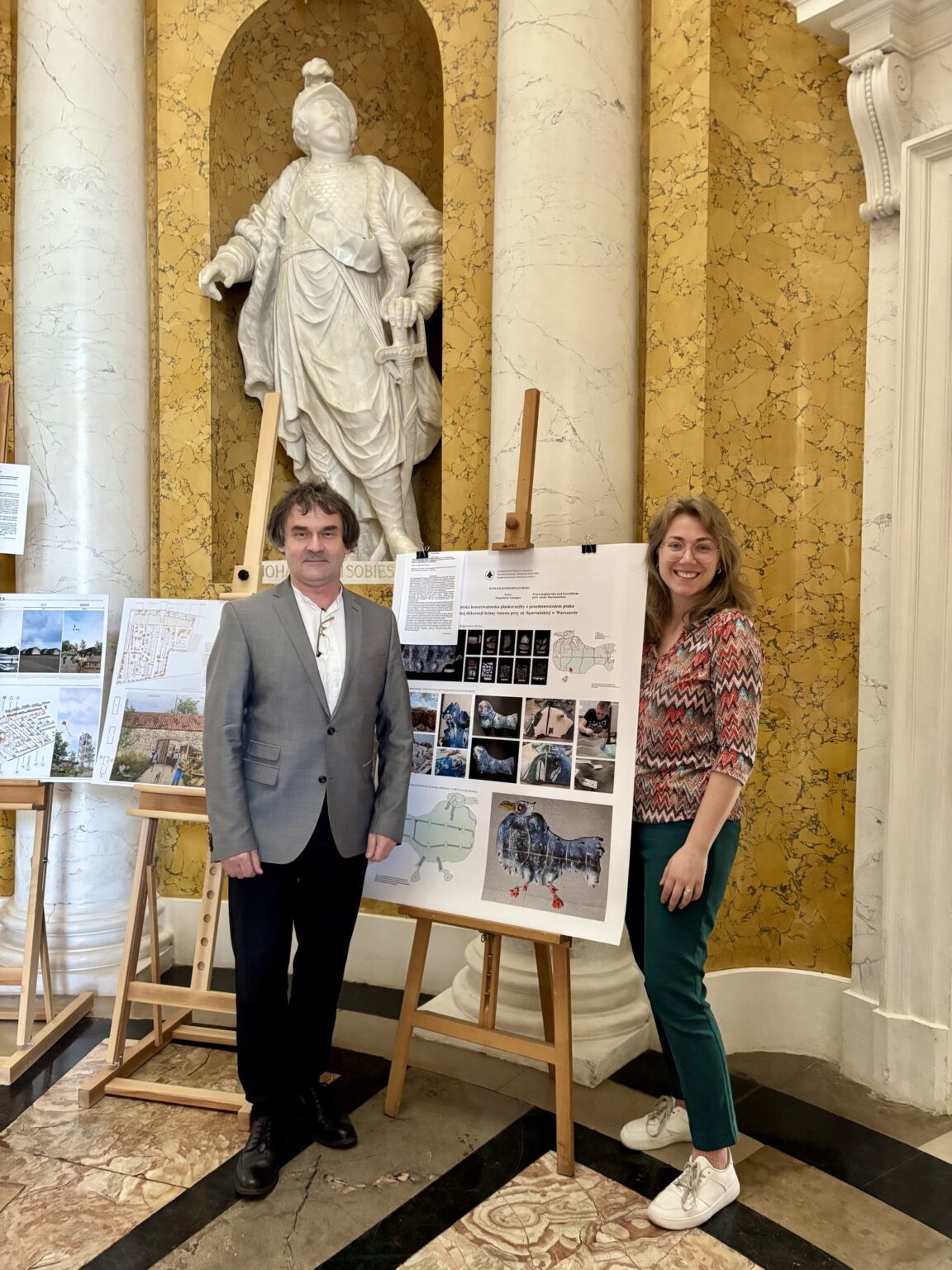 Uczestniczka konkursu i prowadzący projekt stoją przy planszy prezentującej dokumentację konserwatorską ptasiej mozaiki.