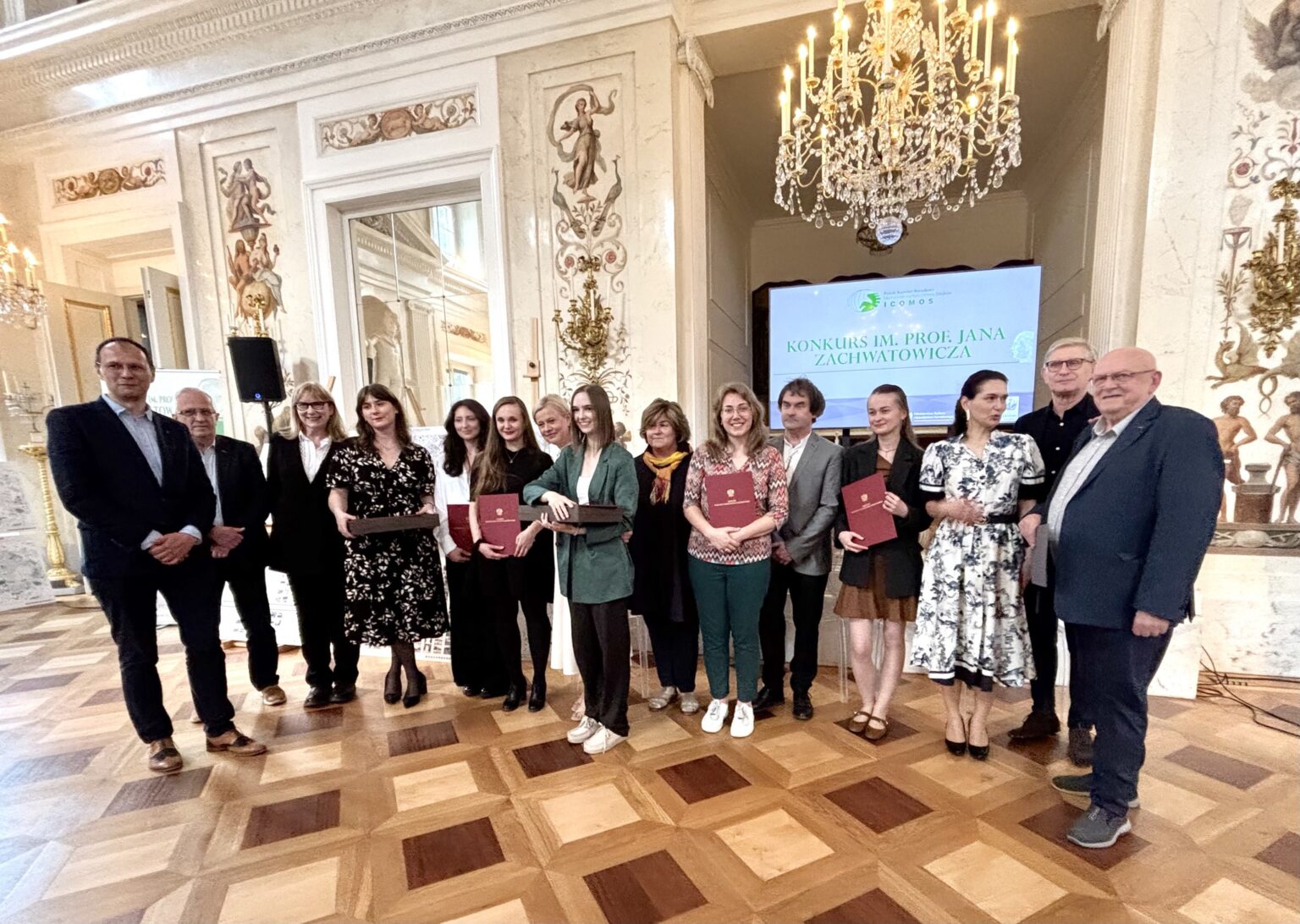 Grupowe zdjęcie laureatów i organizatorów Konkursu im. prof. Jana Zachwatowicza w eleganckim wnętrzu z żyrandolem i ekranem w tle.