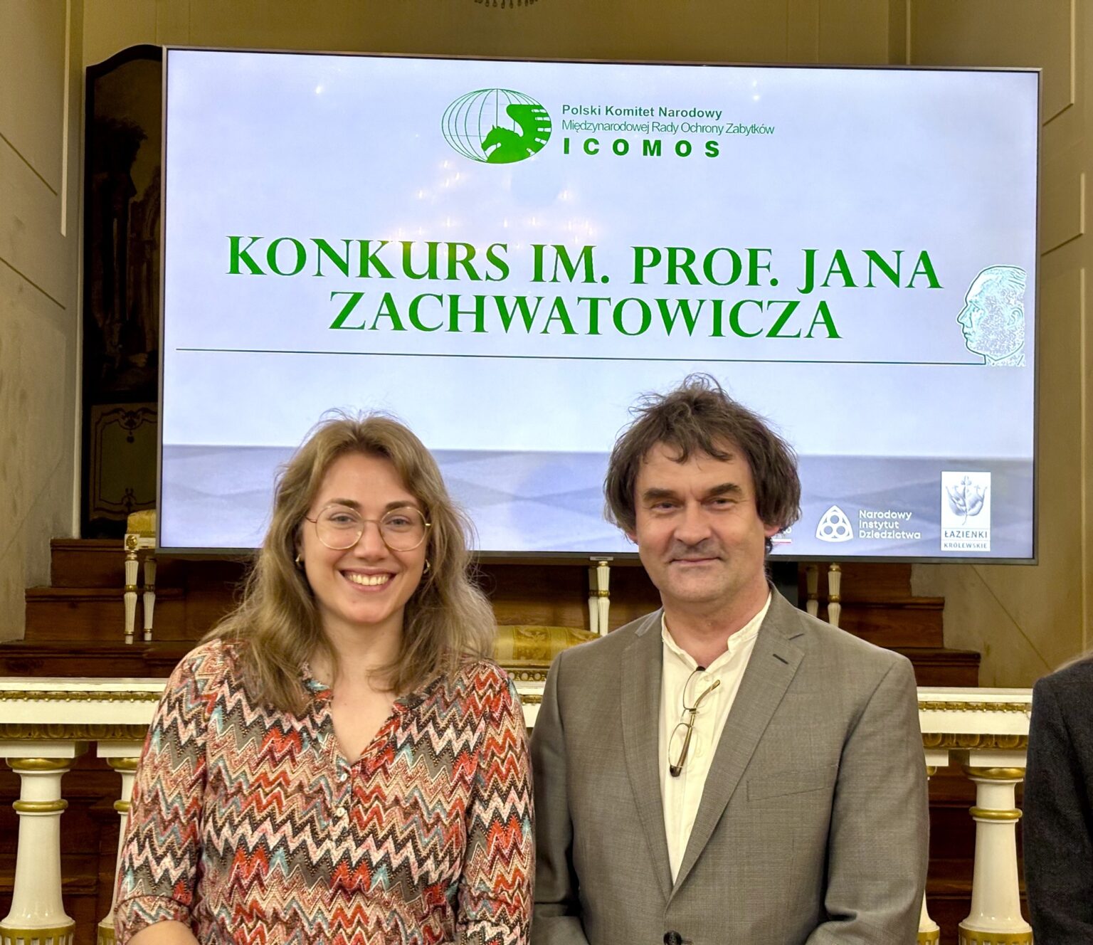 Zbliżenie na uczestników konkursu stojących przed ekranem z logo ICOMOS i nazwą konkursu, trzymających dyplomy i nagrody.