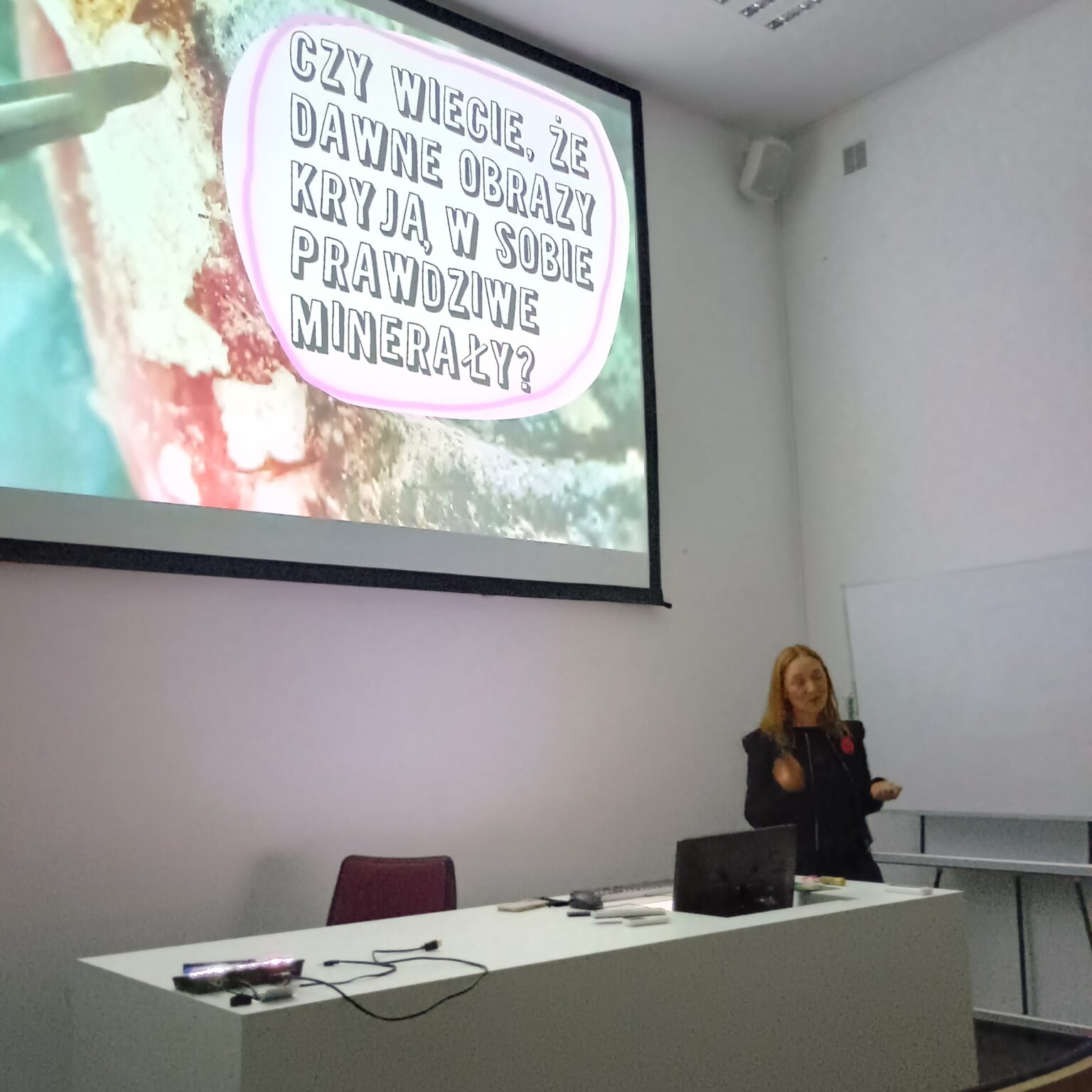 Wykładowczyni stoi obok biurka, gestykulując podczas prezentacji. Na dużym ekranie wyświetlony jest slajd z napisem: „CZY WIECIE, ŻE DAWNE OBRAZY KRYJĄ W SOBIE PRAWDZIWE MINERAŁY?”.