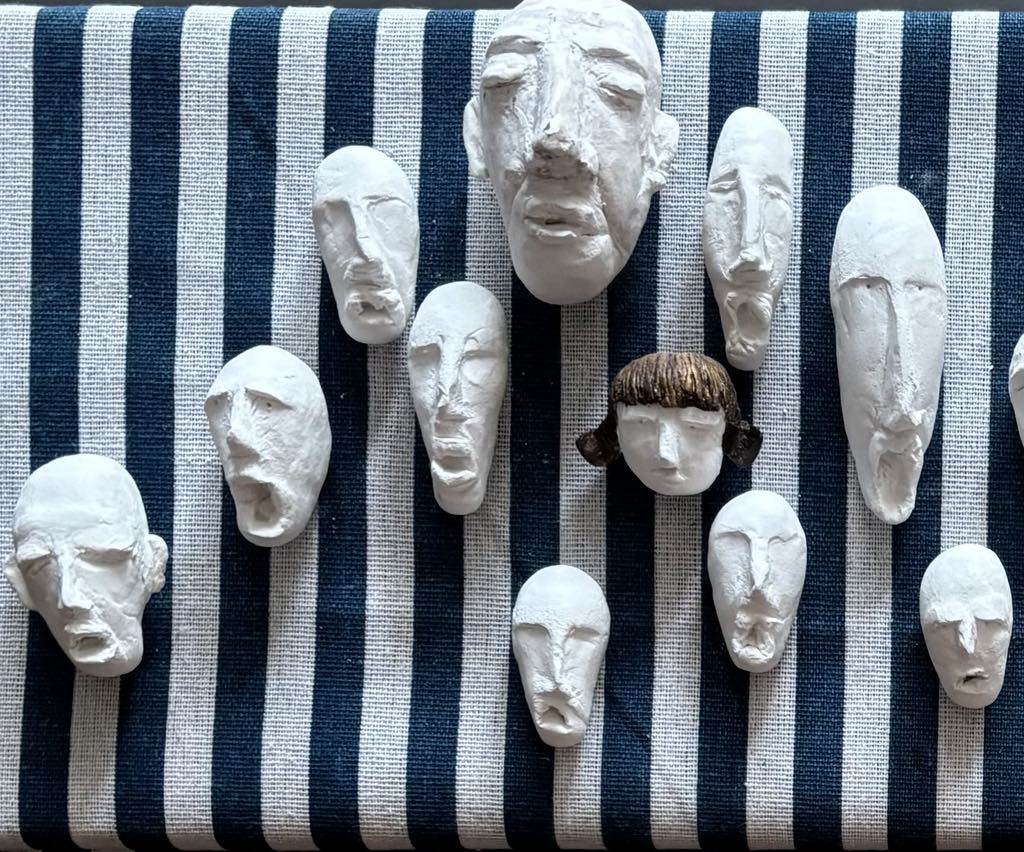 Zbliżenie na fragment pierwszej kompozycji: na tle pionowych pasów znajduje się kilkanaście białych, ekspresyjnych masek-twarzy o różnej wielkości, wśród których wyróżnia się jedna twarz z brązowymi włosami w formie grzywki i dwóch warkoczyków, kontrastująca z pozostałymi białymi.