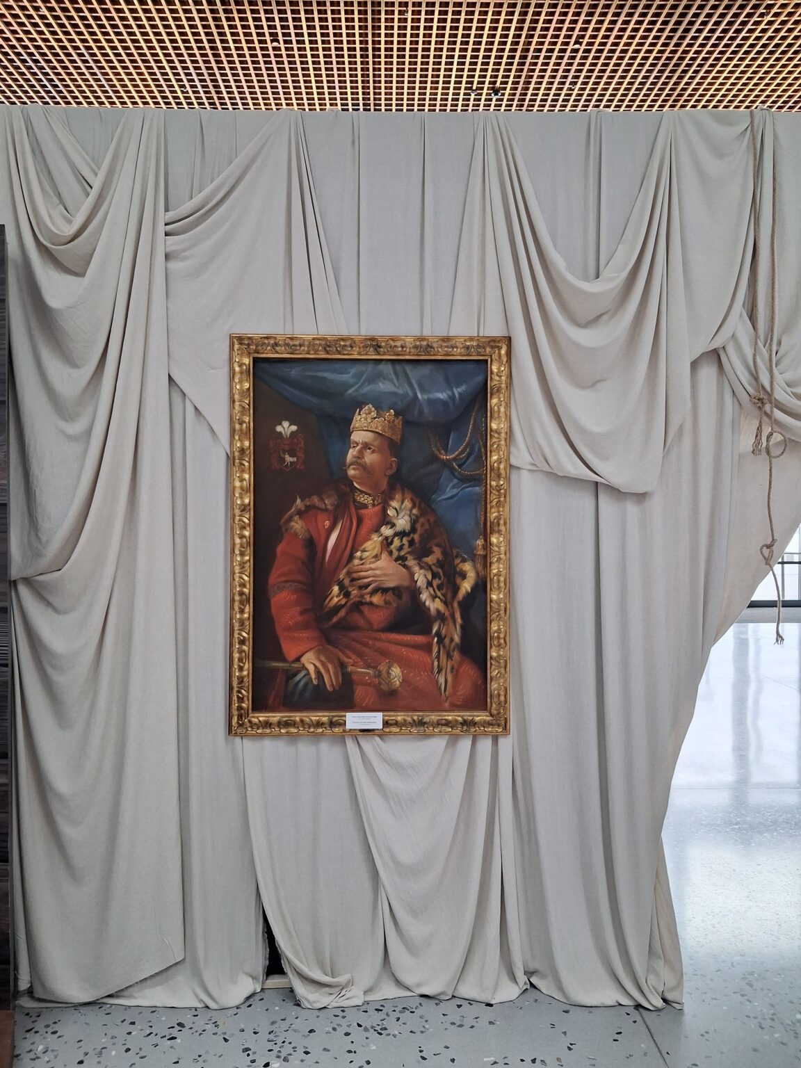 Obraz przedstawiający szlachcica w czerwonym kontuszu z koroną na głowie i płaszczem z futrem, w złotej ramie, zawieszony na tle drapowanych beżowych tkanin.