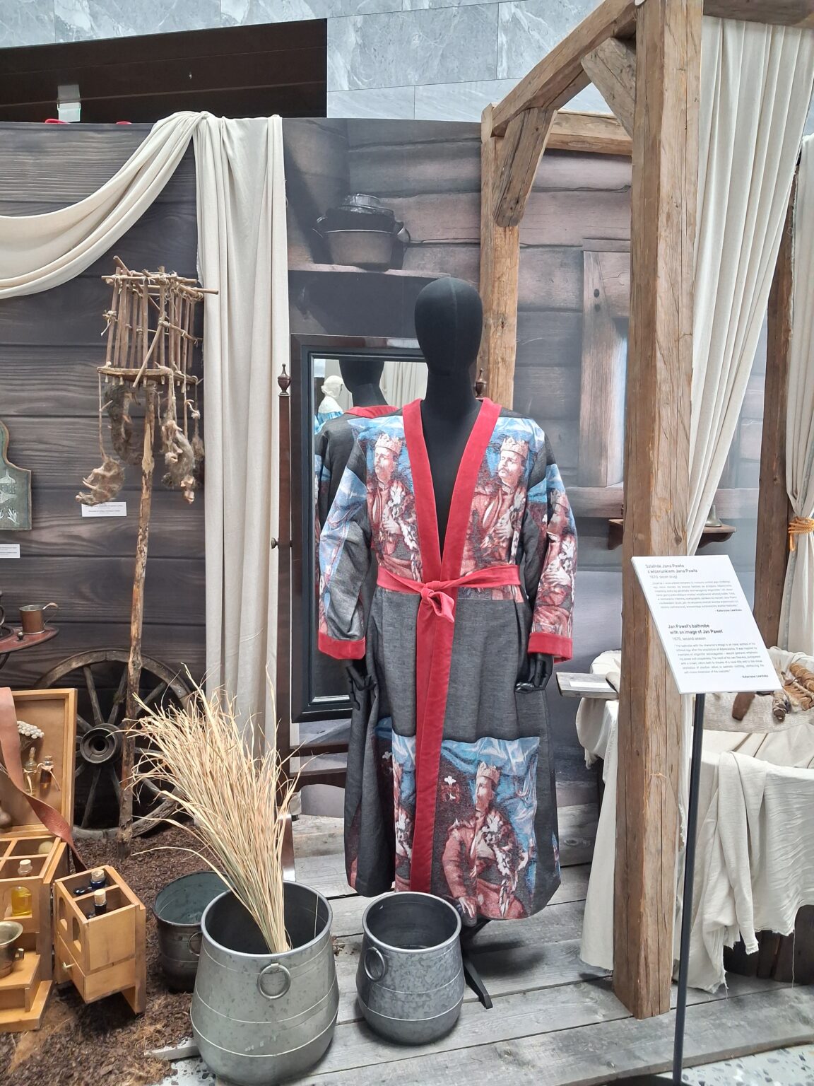 Manekin w kostiumie z serialu 1670 – długi szlafrok ozdobiony nadrukami z wizerunkiem szlachcica w koronie, przepasany czerwonym pasem. Obok widoczne są elementy scenografii wiejskiej: drewniana chata, wiadra i kłosy zboża.