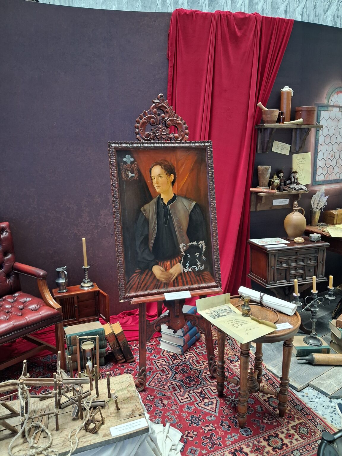 Aranżacja wnętrza stylizowanego na gabinet XVII-wieczny. Na środku stoi portret młodego mężczyzny w ciemnym stroju z herbem i głową dzika, oprawiony w ozdobną ramę. Wokół znajdują się książki, świeczniki, pergaminy i drewniane modele.