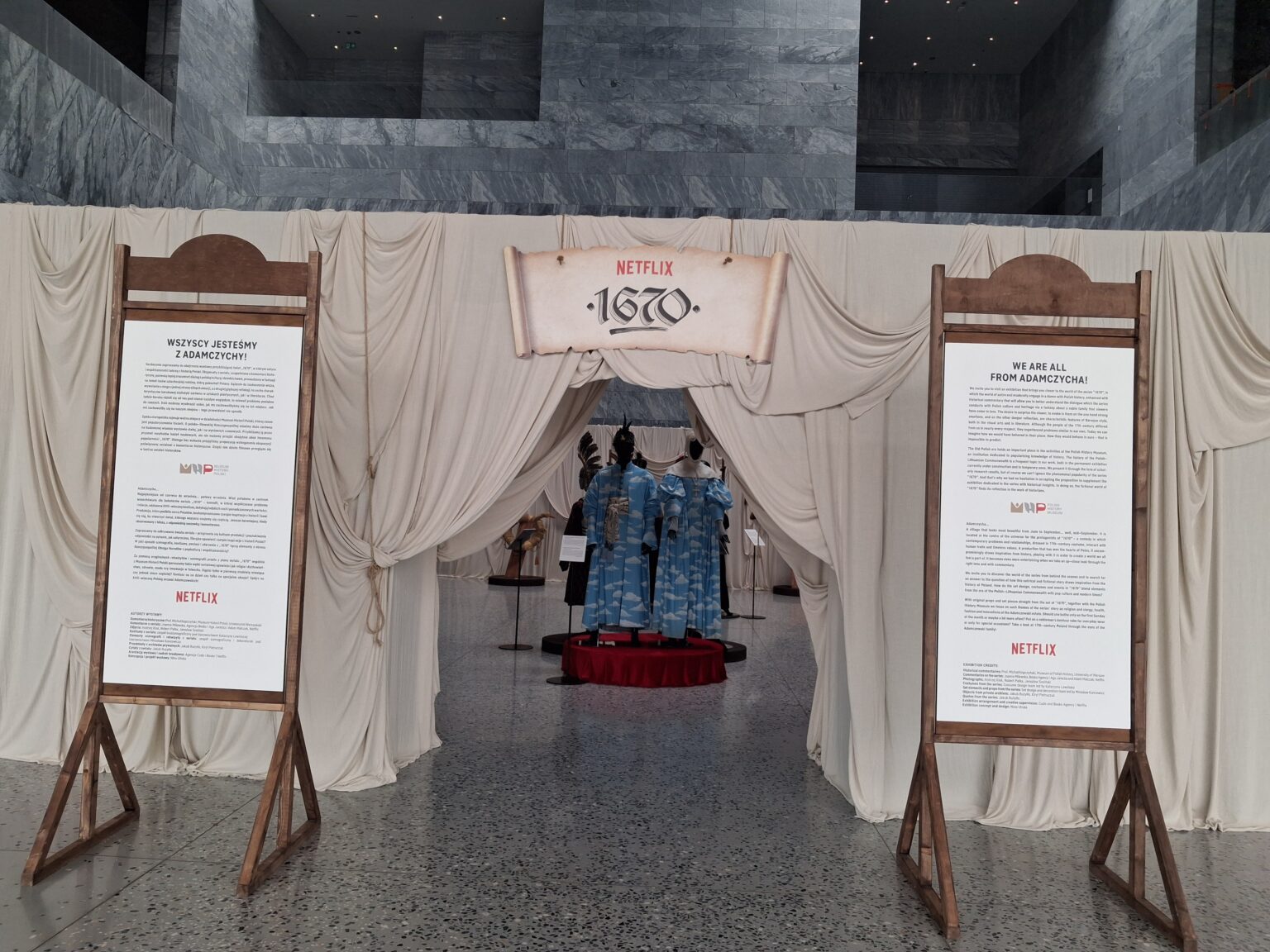 Wejście do wystawy „Wszyscy jesteśmy z Adamczychy” w Muzeum Narodowym w Poznaniu. Na białych płóciennych kotarach zawieszono tablice z opisami po polsku i angielsku, w głębi widoczne są manekiny w kostiumach.