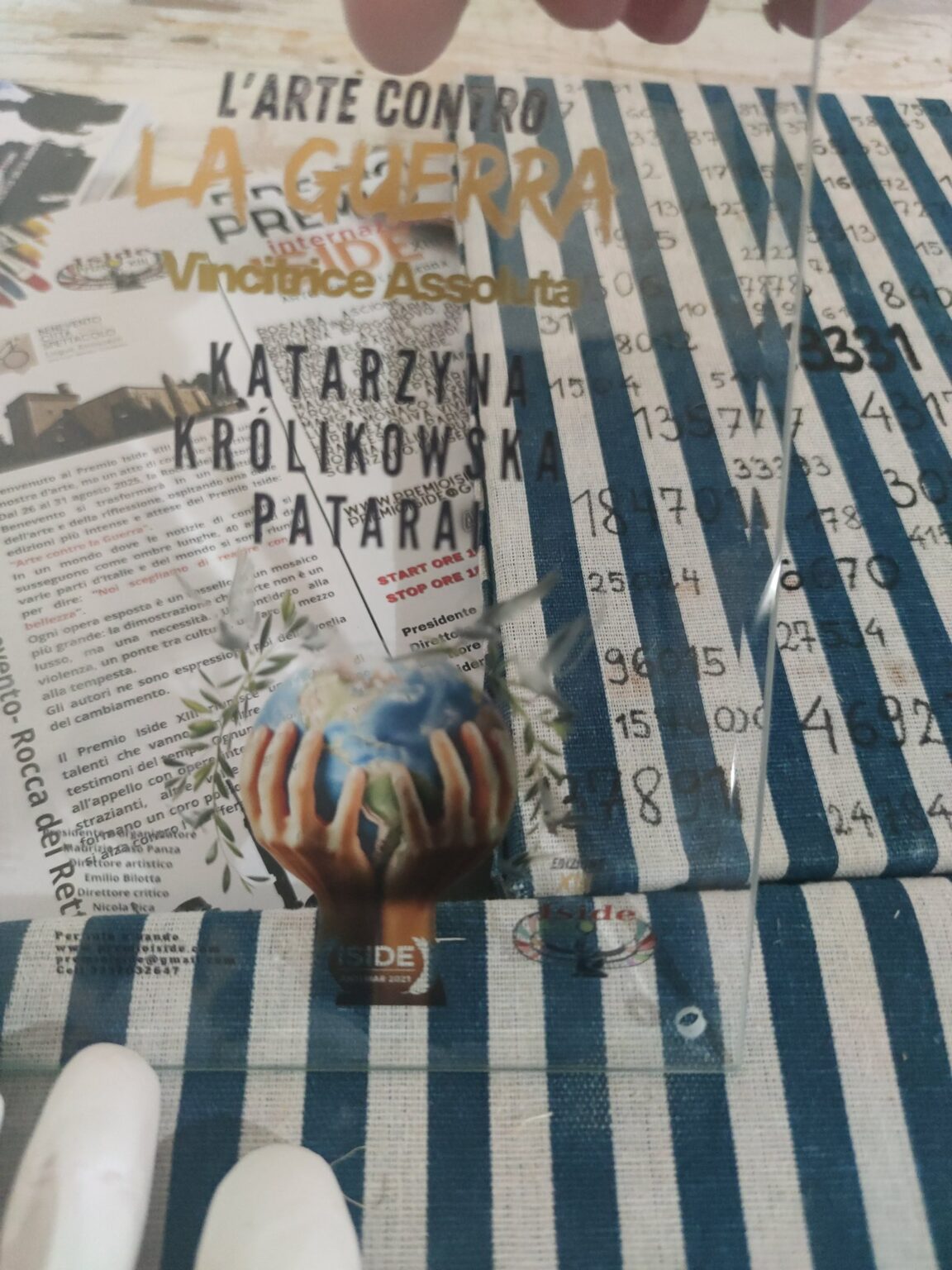 widoczny przez przezroczystą nagrodę z napisem „L’ARTE CONTRO LA GUERRA” i nazwiskiem „Katarzyna Królikowska Pataraci”. W tle dostrzegalne są pasy oraz liczby na płótnie, a na nagrodzie – grafika dłoni trzymających kulę ziemską.