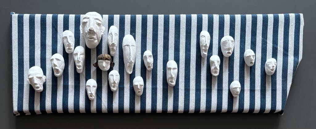 Kompozycja artystyczna na tle pasiaka z pionowych niebiesko-białych pasów. Na powierzchni umieszczono kilkanaście małych, białych rzeźbiarskich masek twarzy o zróżnicowanych, ekspresyjnych wyrazach. Twarze są nierówne, zdeformowane, pełne emocji – sugerują cierpienie i krzyk.