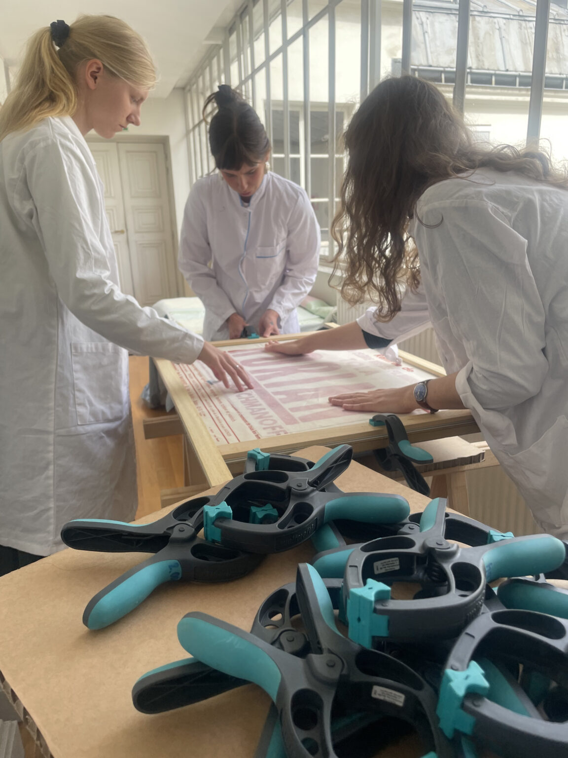 Trzy osoby w białych fartuchach pracują przy dużym stole przy oknie, ostrożnie podnosząc i oglądając stary drukowany plakat.