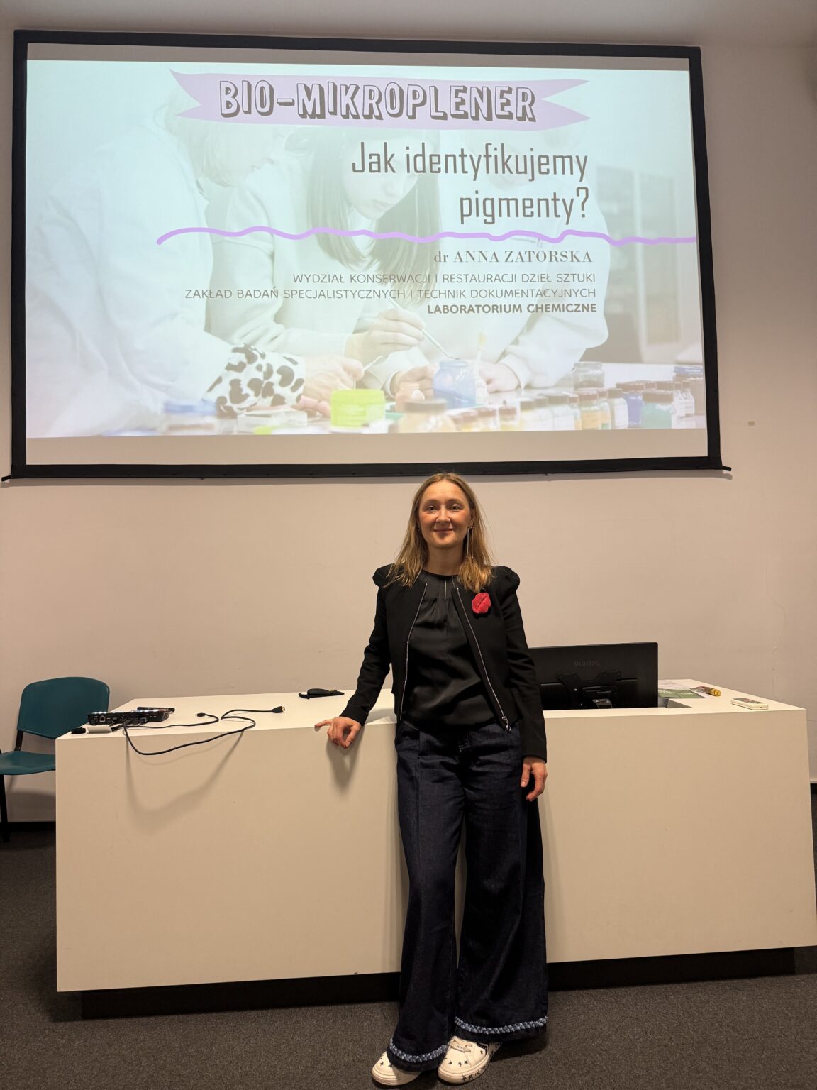 W sali wykładowej kobieta stoi przy biurku i opiera się o nie. Na ekranie za nią wyświetlona jest prezentacja zatytułowana „BIO-MIKROPLENER. Jak identyfikujemy pigmenty?”.