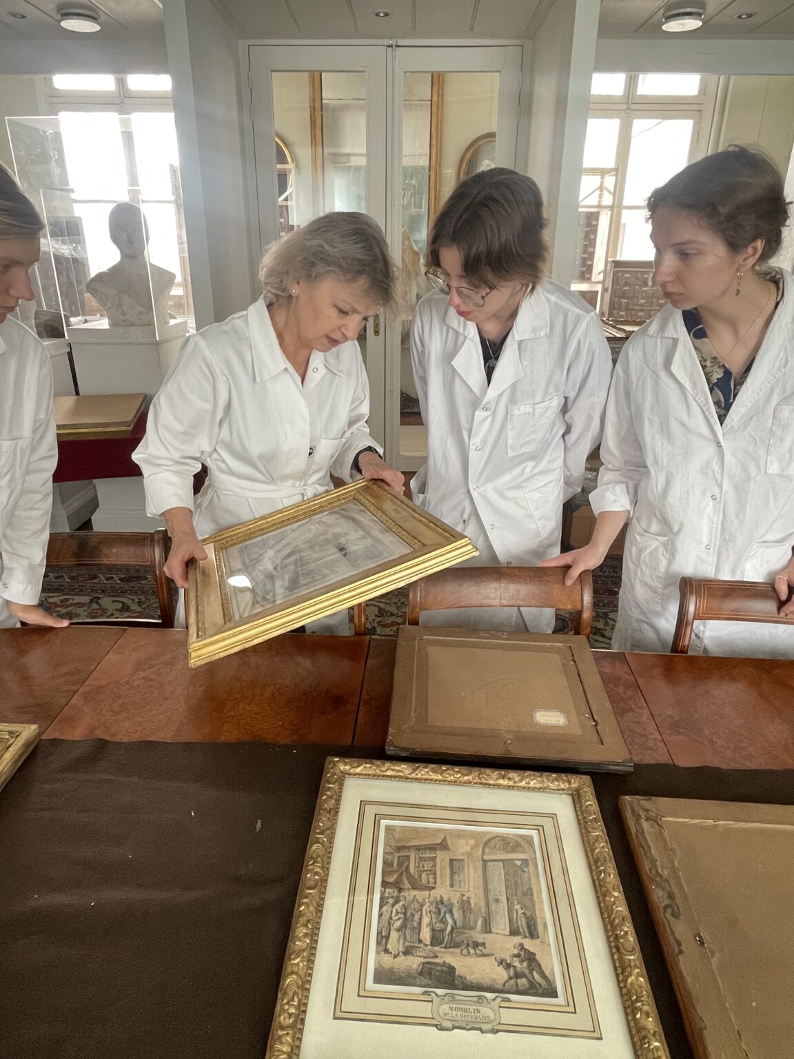 Cztery osoby w białych fartuchach stoją przy stole i analizują obraz w złotej ramie; na stole leży kilka innych oprawionych grafik.