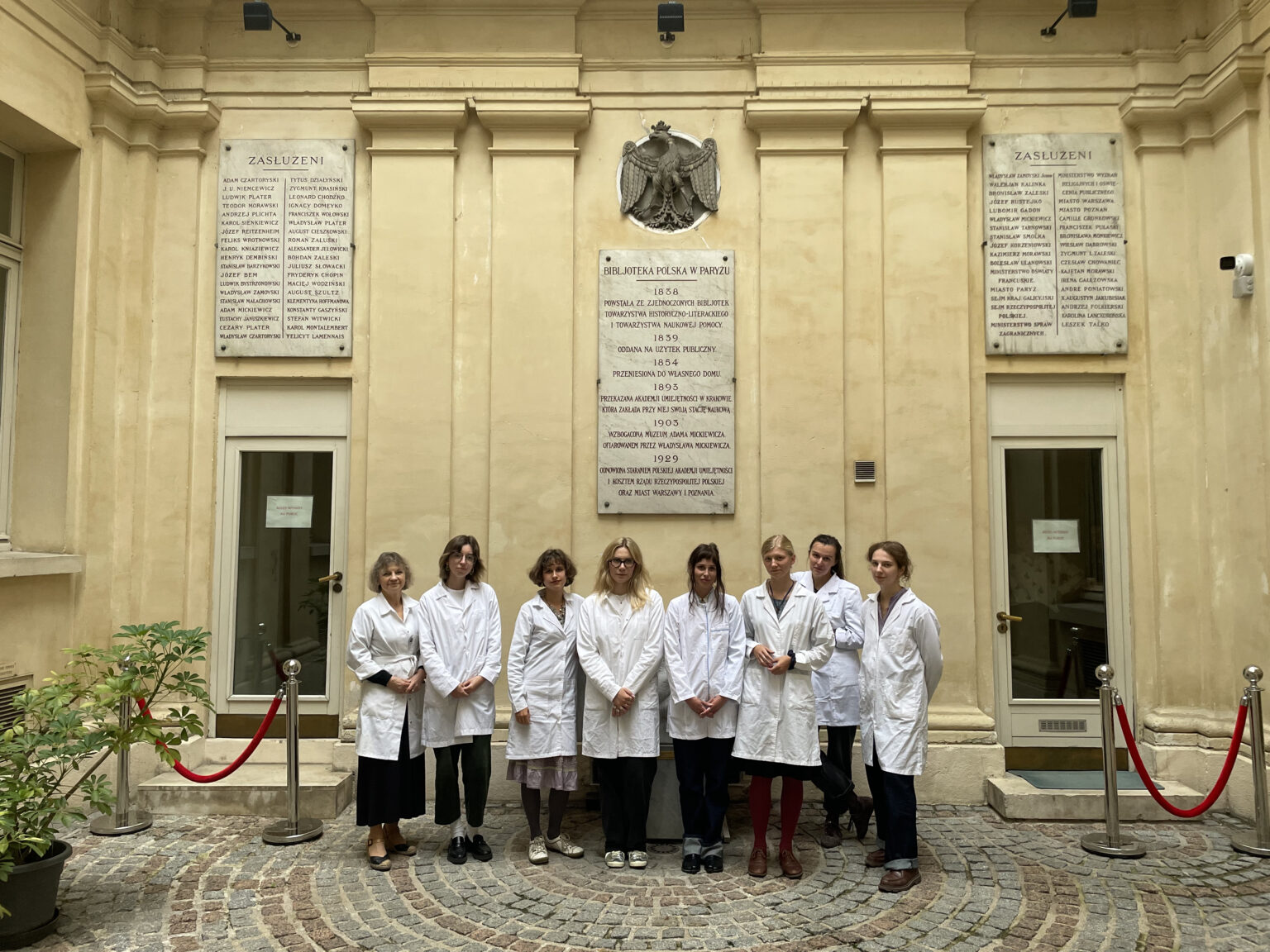 Grupa ośmiu osób w białych fartuchach pozuje wspólnie na dziedzińcu Biblioteki Polskiej w Paryżu, stojąc przed tablicami pamiątkowymi na ścianie.