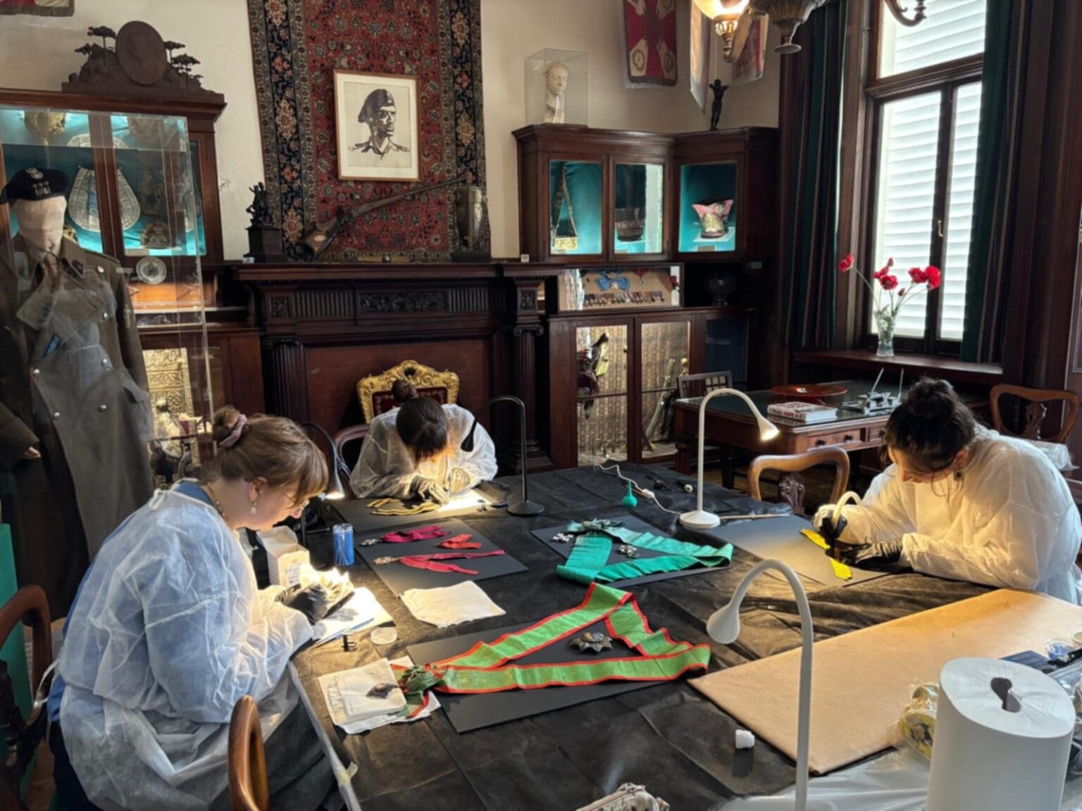 Szeroki plan ukazujący kilka osób pracujących przy stole nad wstęgami, orderami i elementami umundurowania, w historycznym wnętrzu z gablotami i eksponatami muzealnymi.