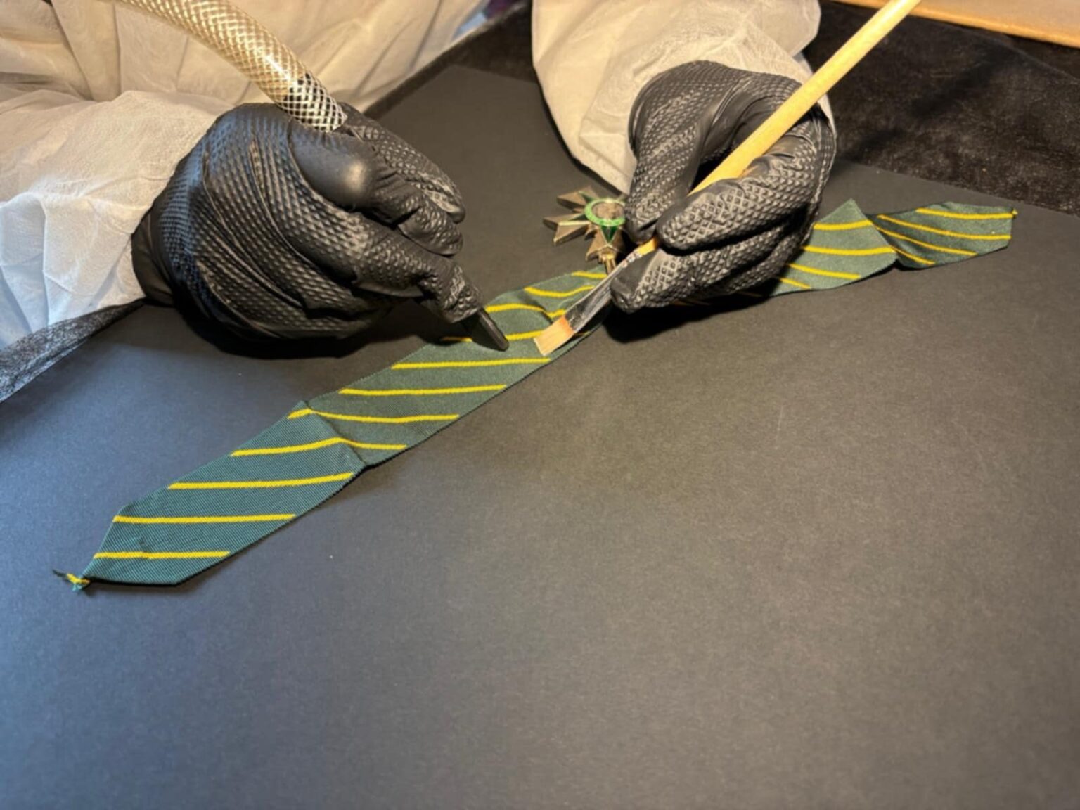 Zbliżenie na dłonie w czarnych rękawiczkach ochronnych, które przy użyciu pędzla i ssaka punktowego oczyszczają zielono-żółtą wstążkę orderową ułożoną na czarnym podłożu.