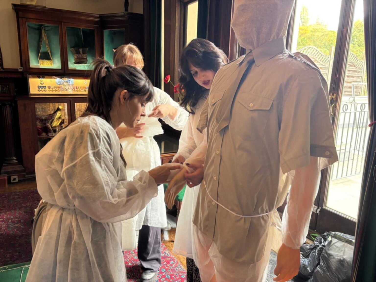 Konserwatorki w fartuchach ochronnych dopasowują rękawicę do manekina ubranego w jasną koszulę wojskową; scena rozgrywa się przy oknie w sali muzealnej.