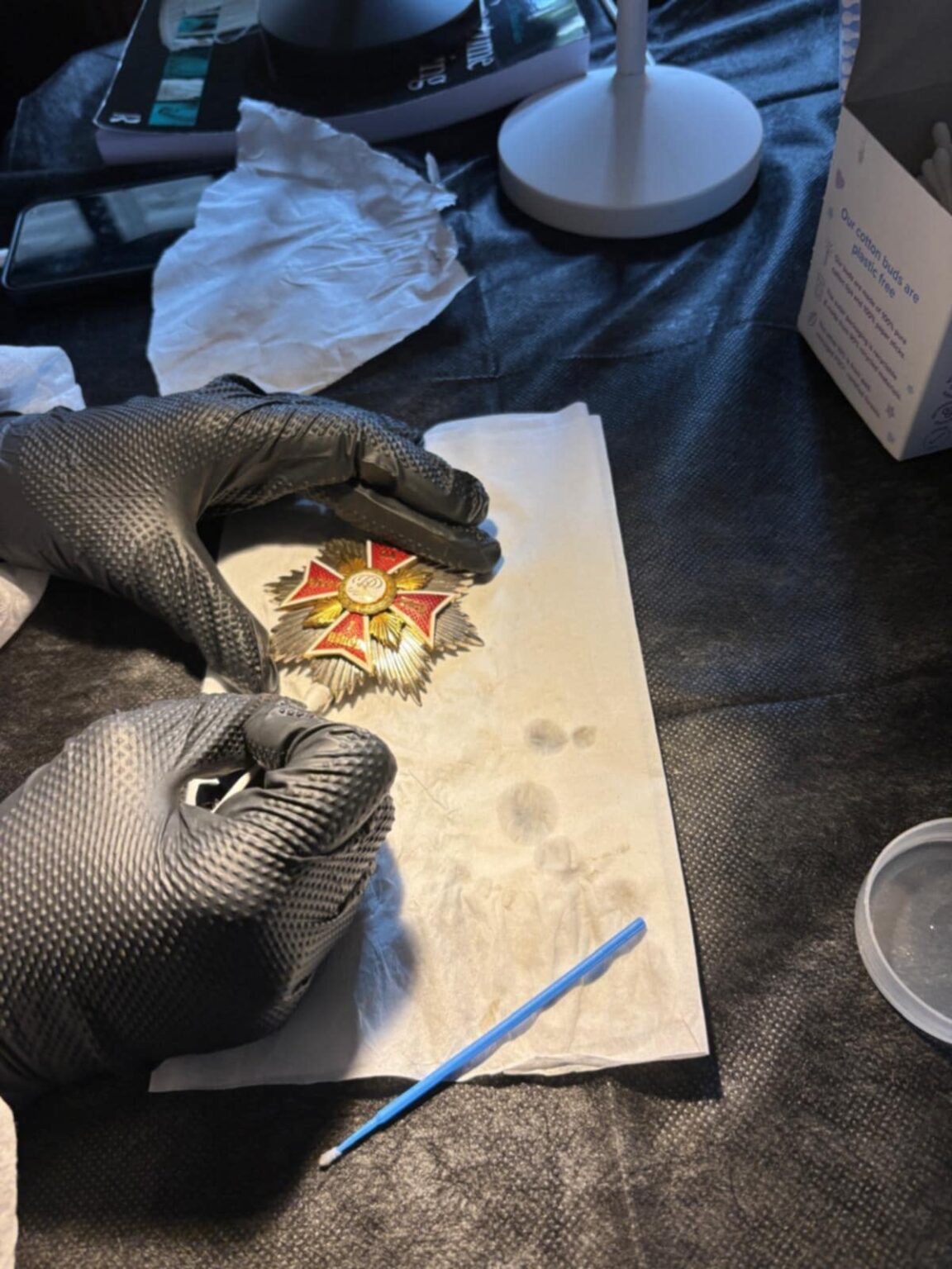 Zbliżenie na dłonie w rękawiczkach podczas mechanicznego oczyszczania metalowego orderu o kształcie gwiazdy, ułożonego na bibułce konserwatorskiej.