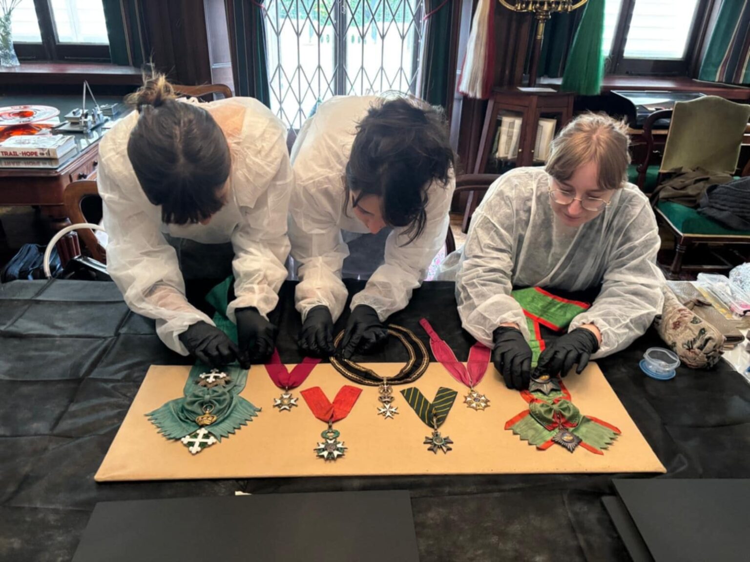 Trzy osoby w białych fartuchach ochronnych pracują przy stole nad zespołem orderów i odznaczeń wojskowych, precyzyjnie porządkując wstęgi i elementy metalowe.