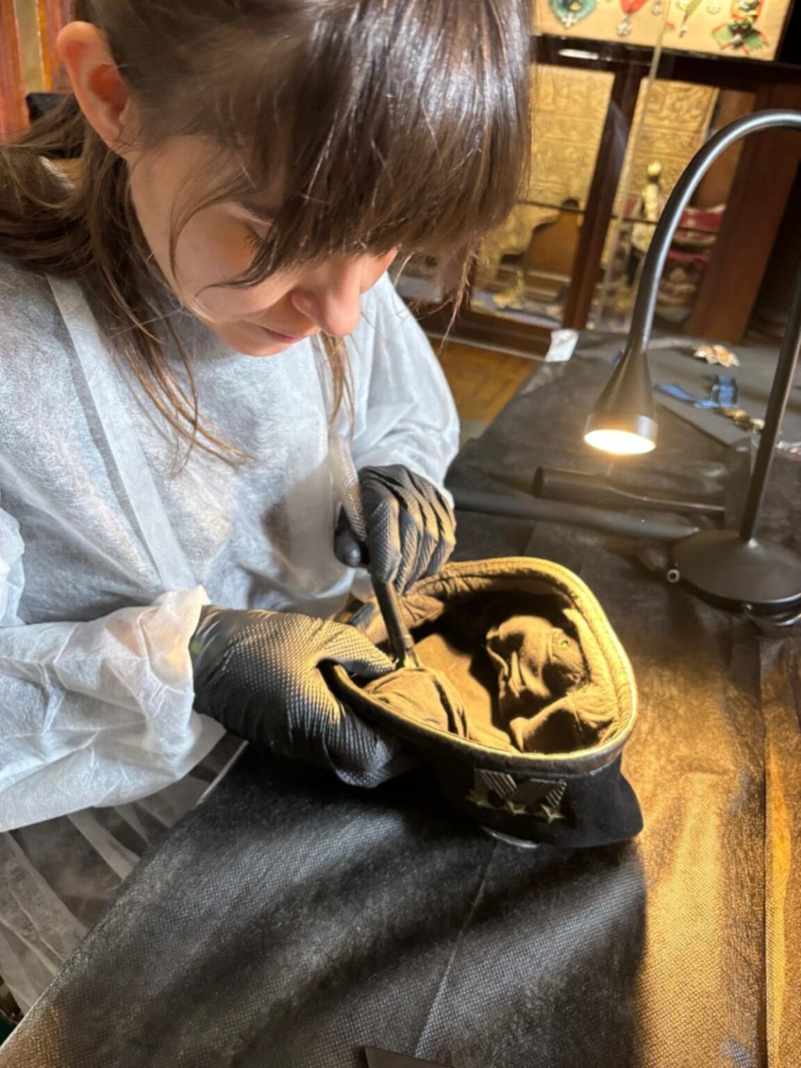 Konserwatorka w rękawiczkach i fartuchu ochronnym wykonuje precyzyjne czynności przy wnętrzu wojskowej czapki, oświetlonej lampą stanowiskową.